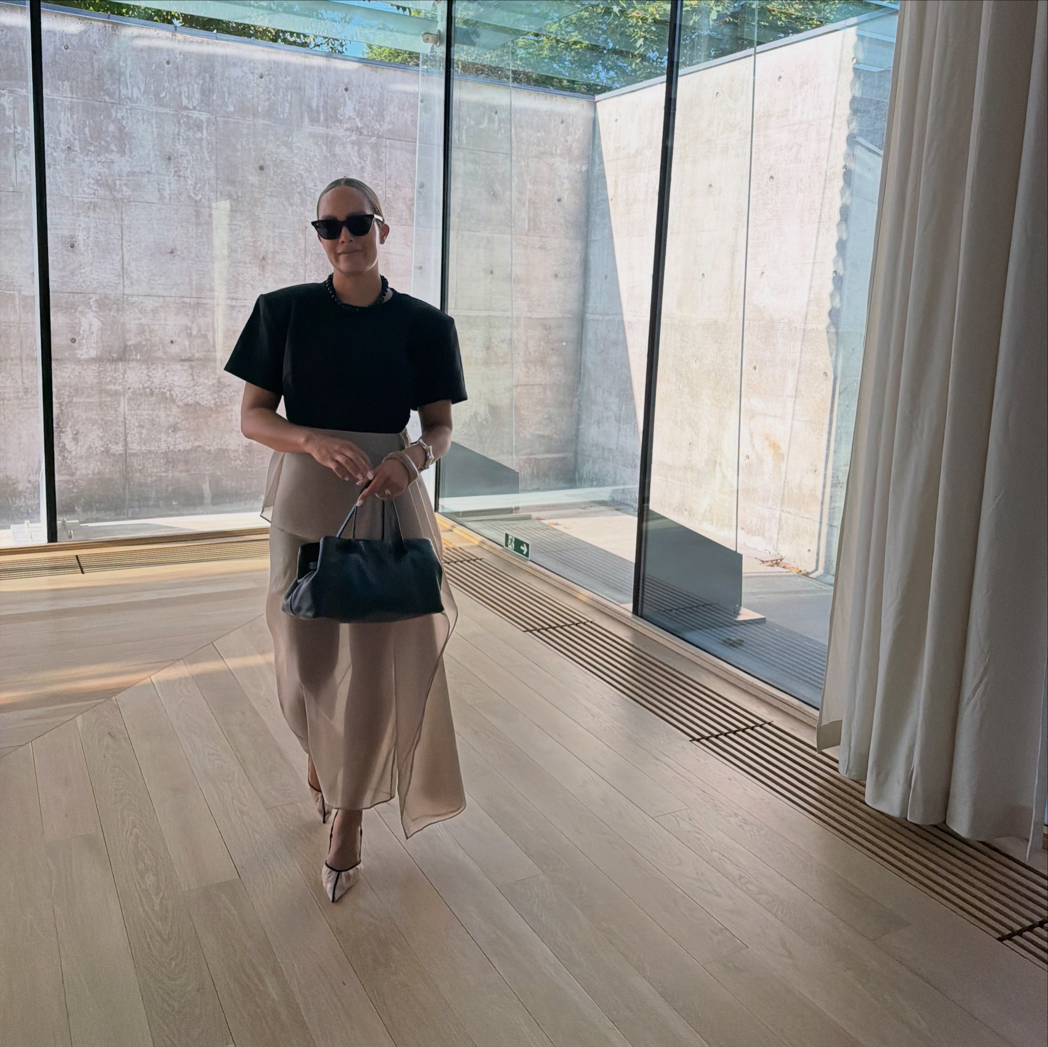 Nicole wearing a black top and flowing beige skirt, carrying a black handbag beside tall glass windows